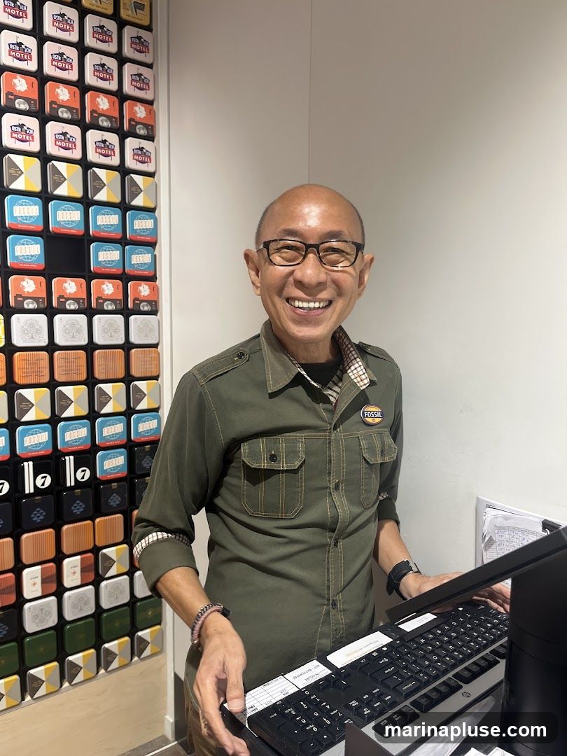 A smiling man wearing glasses and a green Fossil shirt stands behind a desk with a keyboard. To his left is a wall display featuring numerous small, square items with various designs, many of which appear to be tins or boxes. The Fossil logo is visible on his shirt pocket.
