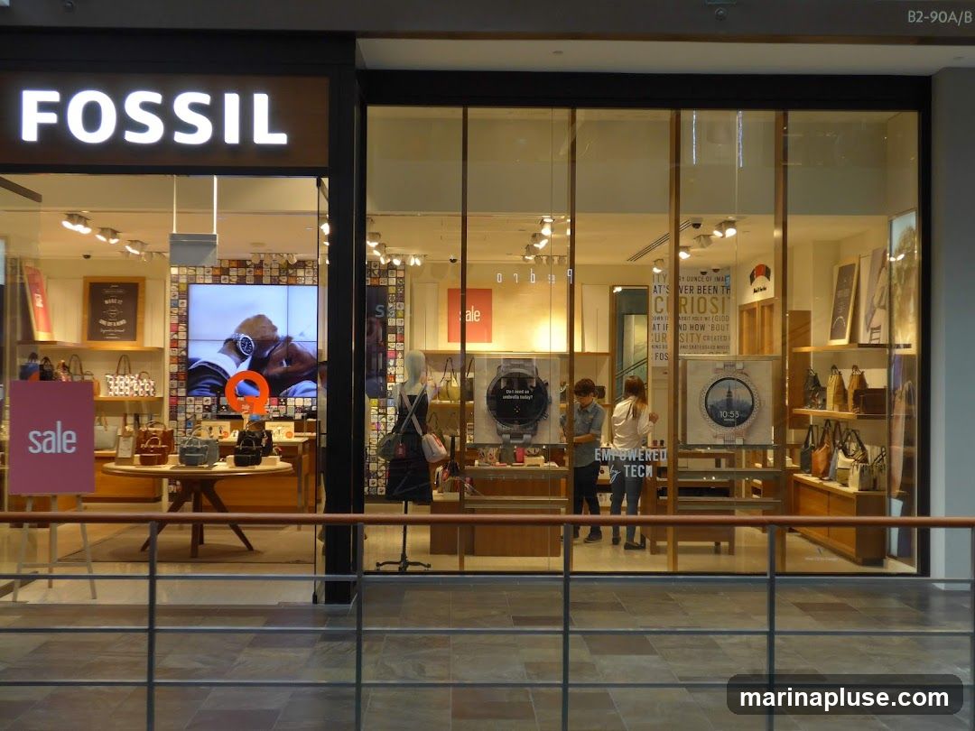 The exterior of a Fossil store with a large illuminated "FOSSIL" sign above the entrance. The store features large glass windows displaying watches, bags, and other accessories. A mannequin is visible inside, and a "sale" sign is placed outside the window.