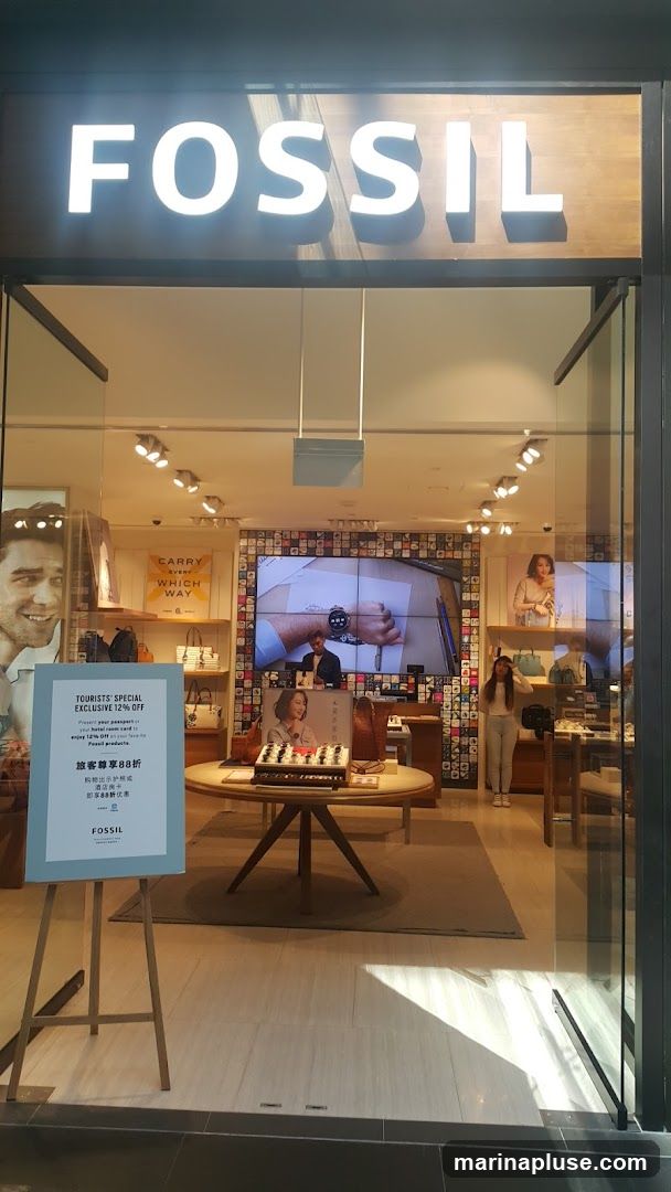 The exterior of a Fossil store with the brand name prominently displayed in large white letters above the entrance. Through the glass doors, the interior of the store is visible, showcasing displays of watches, handbags, and promotional screens. A sign near the entrance advertises a "Tourists Special Exclusive 12% Off" and also displays text in Chinese.