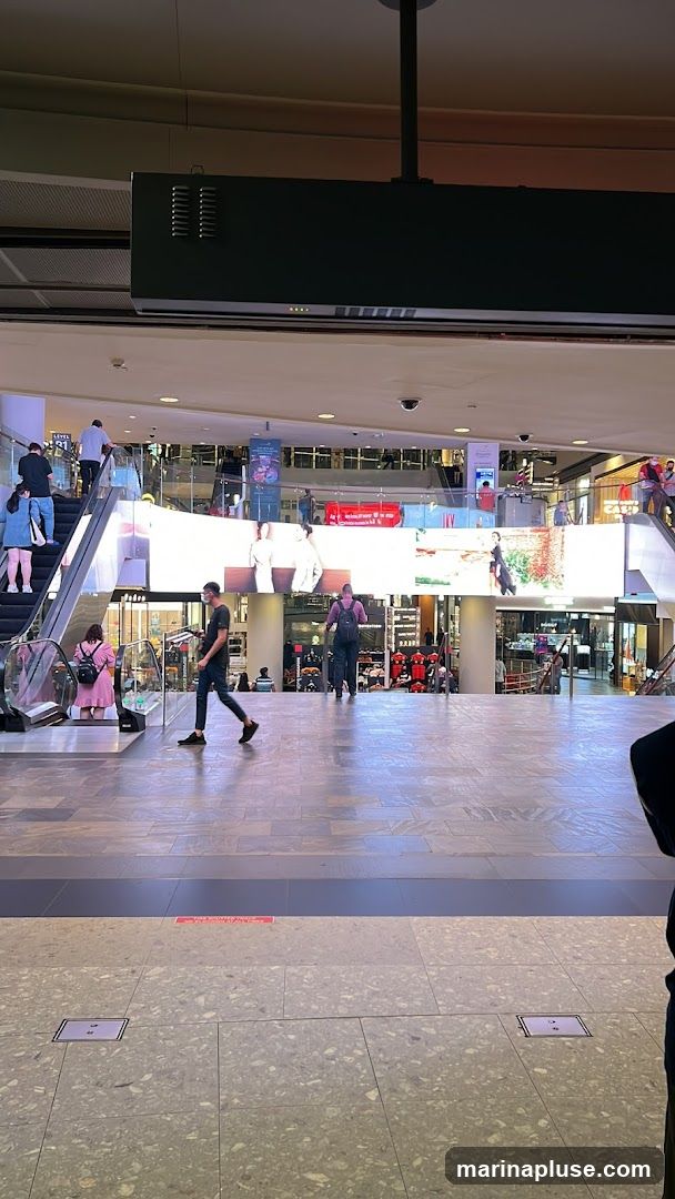 A wide shot of a bustling shopping mall interior. Escalators are visible on the left and right sides, with people using them. A large, illuminated digital screen spans across the middle of the frame, displaying various images and advertisements. The floor is tiled with a speckled pattern. Several shops and kiosks are visible in the background, with some shoppers browsing.
