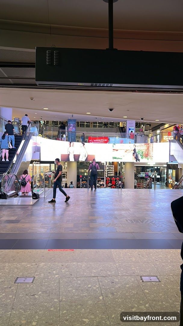 A wide shot of a bustling shopping mall interior. Escalators are visible on the left and right sides, with people using them. A large, illuminated digital screen spans across the middle of the frame, displaying various images and advertisements. The floor is tiled with a speckled pattern. Several shops and kiosks are visible in the background, with some shoppers browsing.
