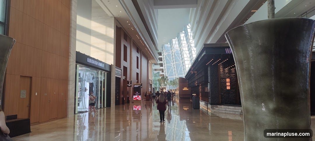 A wide, well-lit interior space with a high ceiling and a glass facade on one side, revealing a modern architectural structure. The floor is highly reflective, mirroring the surroundings. On the left, a store with the sign "RICHARD MILLE" is visible, featuring a glass storefront and mannequins. Further down, another store has a sign that reads "Hair Studio". On the right, a dark-toned establishment with a sign that starts with "WA" is present. Large, abstract sculptures are placed in the foreground. Several people are walking through the space.