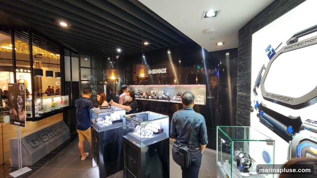 The interior of a G-SHOCK store, featuring display cases filled with watches. A large G-SHOCK advertisement is visible on the right wall. Several customers and staff members are present.
