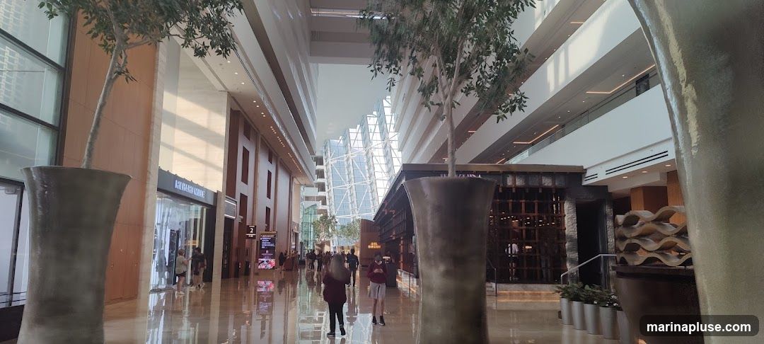 A wide shot of a modern indoor atrium with multiple levels. Large potted trees are placed strategically throughout the space. On the left, a storefront with the name "RICHARD KAREN" is visible. Further down the corridor, a sign indicates "2". On the right, a restaurant or bar area with a distinctive wooden lattice facade is present. The atrium features a reflective floor and a large, angular glass structure in the distance. Several people are walking through the space.