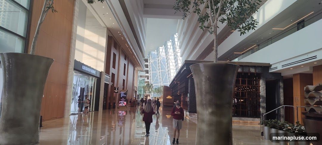 A wide shot of a modern indoor atrium with a high ceiling and large windows. The atrium features polished floors, wooden paneling on the walls, and large decorative planters with trees. Several shops are visible along the sides, including one with the signage "RICHARD KNI..." and another with "BATHROOM". People are walking through the space. The architecture includes geometric patterns and a sense of spaciousness.