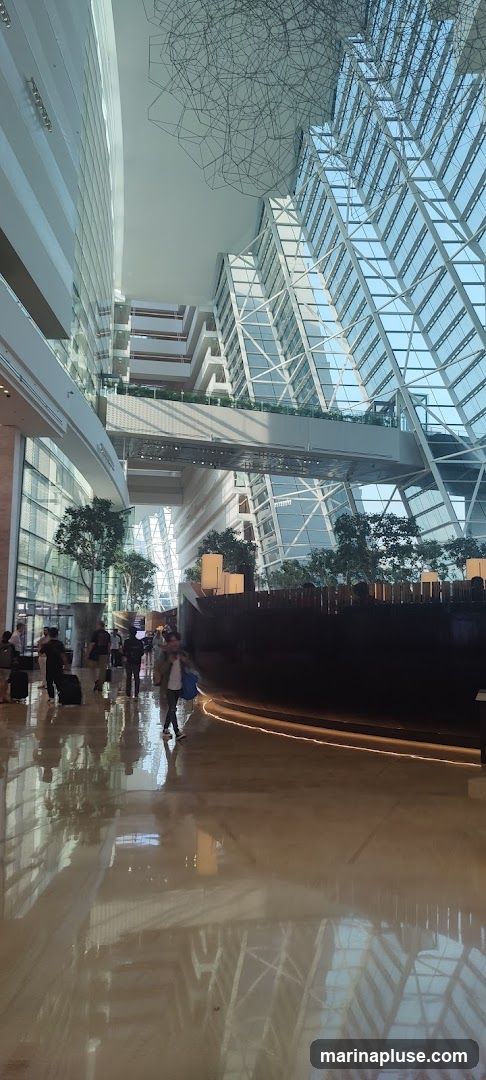 A wide shot of a spacious and modern hotel lobby with a high ceiling and a glass facade. People are seen walking with luggage, suggesting a busy travel hub. The architecture features multiple levels connected by walkways and a striking geometric art installation hanging from the ceiling. Lush indoor plants are strategically placed throughout the space, adding a touch of nature to the contemporary design. The polished floor reflects the light and the surrounding structures, enhancing the sense of grandeur.