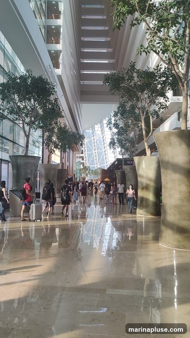 A wide, sunlit indoor walkway with a polished floor reflecting the surroundings. Large potted trees line the walkway, and people are seen walking in both directions. The architecture features multiple levels with glass facades and a distinctive geometric structure in the distance.