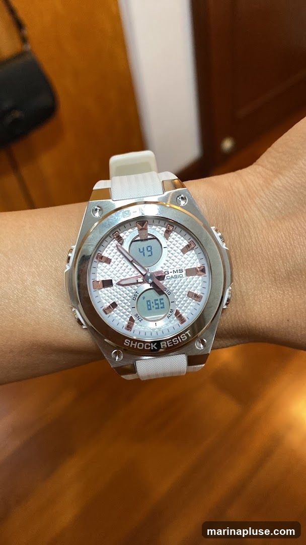 A close-up shot of a woman's wrist wearing a silver and white Casio G-MS watch. The watch has a white textured dial with rose gold accents and hands. It features two digital displays, one showing '49' and the other '8:55'. The watch band is white and the text 'SHOCK RESIST' is visible on the bezel. A black purse is blurred in the background.