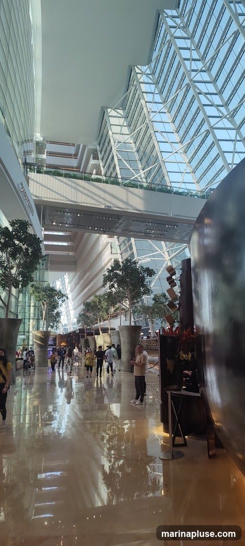 A wide shot of a modern atrium with a high, glass-paneled ceiling and multiple levels of balconies. Large, stylized planters with trees are placed throughout the space, and the polished floor reflects the architecture and greenery. Several people are walking through the atrium, and some are gathered near what appears to be a bar or service area on the right. The overall impression is one of a spacious, contemporary indoor environment.