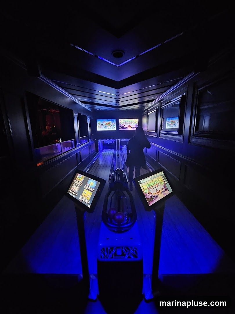 An interior view of a dimly lit bowling alley with arcade-style games. The lanes are illuminated with blue LED lights. There are screens displaying game graphics at the start of each lane, and larger screens above the lanes showing bowling scores or game visuals. A person is visible walking down one of the lanes.