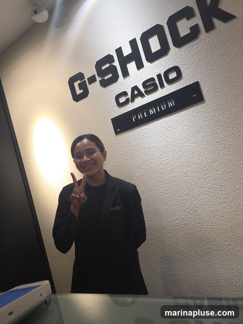 A woman in a black blazer stands in front of a wall with large G-SHOCK CASIO PREMIUM lettering. She is smiling and holding up two fingers in a peace sign.