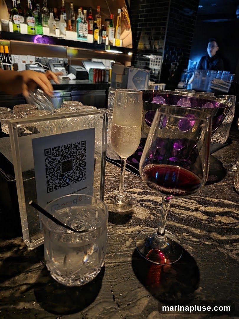 A close-up view of a bar counter with several drinks and glasses. In the foreground, a glass of clear liquid with ice and a black straw sits on a dark, speckled countertop. To its right, a champagne flute with a small amount of bubbly liquid is visible, followed by a wine glass filled with red wine. Behind these, a decorative purple bowl and more glasses are present. The background shows a bar area with shelves stocked with various bottles of alcohol and a bartender in the distance. A QR code is displayed on a clear stand in the foreground.