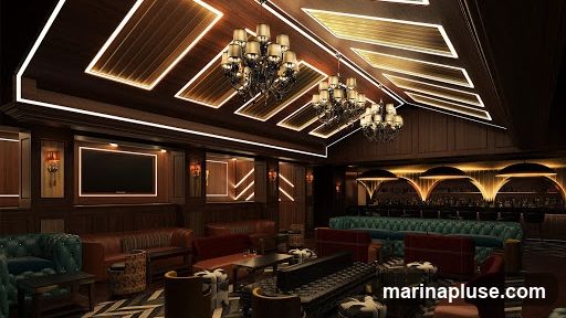 A dimly lit, opulent lounge with dark wood paneling, plush seating in shades of green and red, and ornate chandeliers. The ceiling features modern LED strip lighting integrated into wooden panels. A long bar with illuminated arched backlighting is visible in the background, stocked with bottles. Several flat-screen televisions are mounted on the walls. The overall ambiance suggests a high-end establishment for relaxation and socializing.