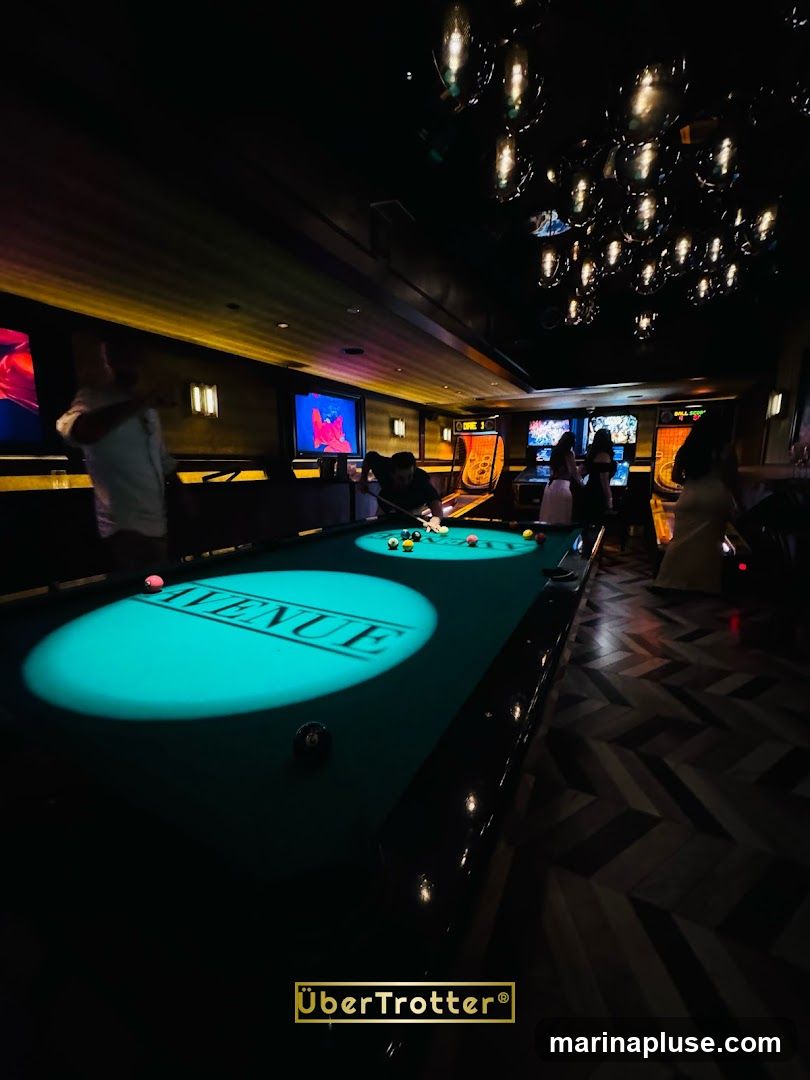 A dimly lit billiard hall with a pool table in the foreground. The word "AVENUE" is projected onto the pool table. Several people are visible, some playing pool and others standing. In the background, there are arcade games and screens displaying colorful images. The ceiling features a cluster of decorative lights. The floor has a herringbone pattern. A watermark for "UberTrotter" is present at the bottom.