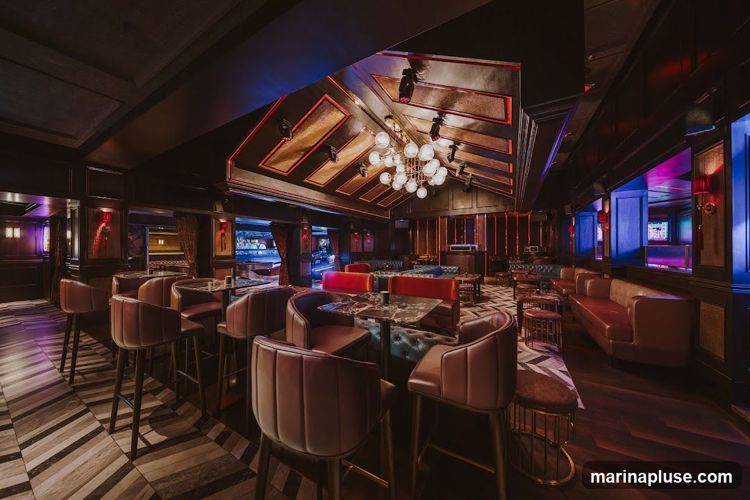A dimly lit, upscale bar interior with plush seating, marble-topped tables, and a striking chandelier. The decor features dark wood paneling, red and blue accent lighting, and a patterned floor. Bar stools are arranged around tables, and lounge seating is visible in the background.