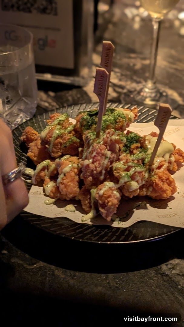 A plate of crispy fried chicken bites drizzled with a creamy sauce and sprinkled with green herbs. The bites are served on a black plate with patterned paper underneath, featuring the word "AVENUE" repeated. Wooden skewers with "AVENUE" branding are stuck into the chicken. A glass of white wine and a partially visible glass with a logo are in the background.