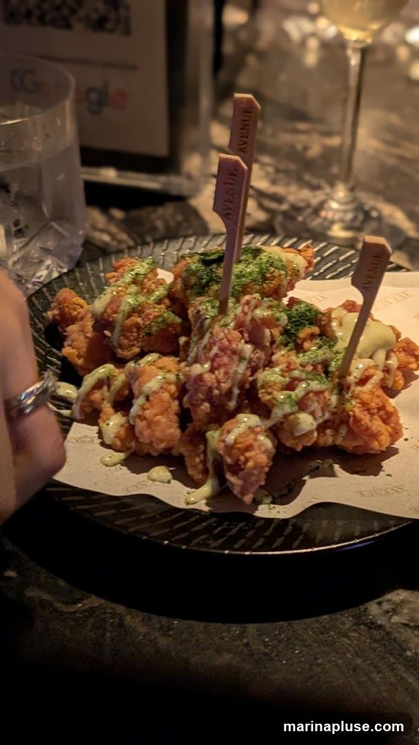 A plate of crispy fried chicken bites drizzled with a creamy sauce and sprinkled with green herbs. The bites are served on a black plate with patterned paper underneath, featuring the word "AVENUE" repeated. Wooden skewers with "AVENUE" branding are stuck into the chicken. A glass of white wine and a partially visible glass with a logo are in the background.