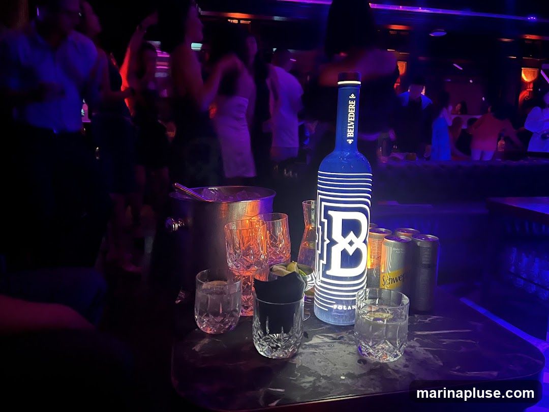 A table in a dimly lit nightclub setting with a prominent blue Belvedere vodka bottle, several cans of Schweppes, and ice bucket with glasses. People are blurred in the background, dancing.