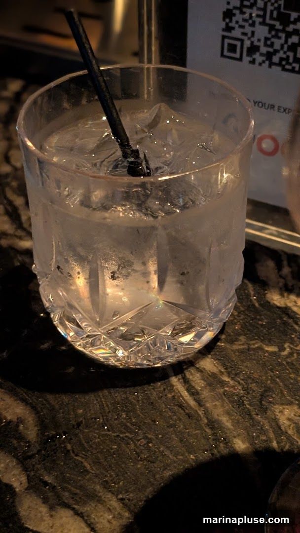 A close-up shot of a clear glass filled with ice and a clear liquid, with a black straw. The glass has a decorative, faceted design at the bottom. The drink is condensation-covered. In the background, a QR code and some text are visible on a white card, along with a hint of a metallic surface. The drink sits on a dark, speckled countertop.