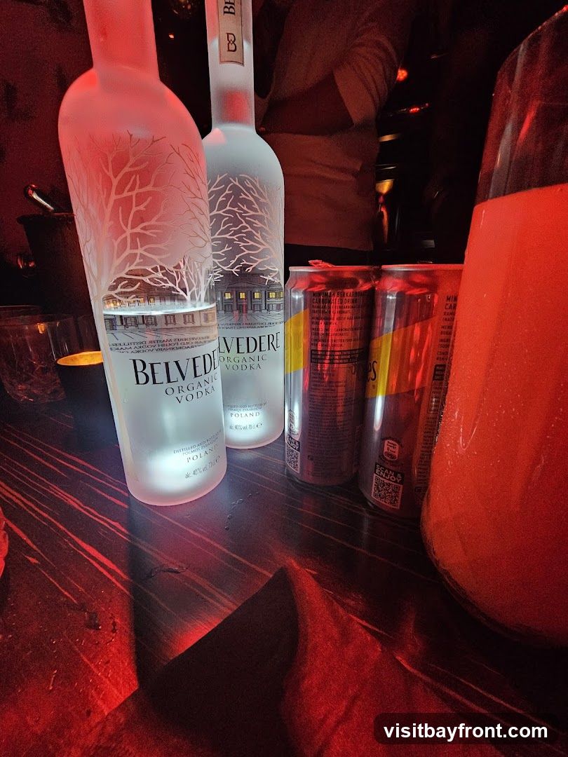 Two bottles of Belvedere Organic Vodka and two cans of Red Bull are on a dark wooden table, illuminated by red lighting. A large glass pitcher filled with a red beverage is to the right.
