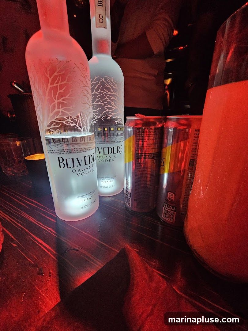 Two bottles of Belvedere Organic Vodka and two cans of Red Bull are on a dark wooden table, illuminated by red lighting. A large glass pitcher filled with a red beverage is to the right.