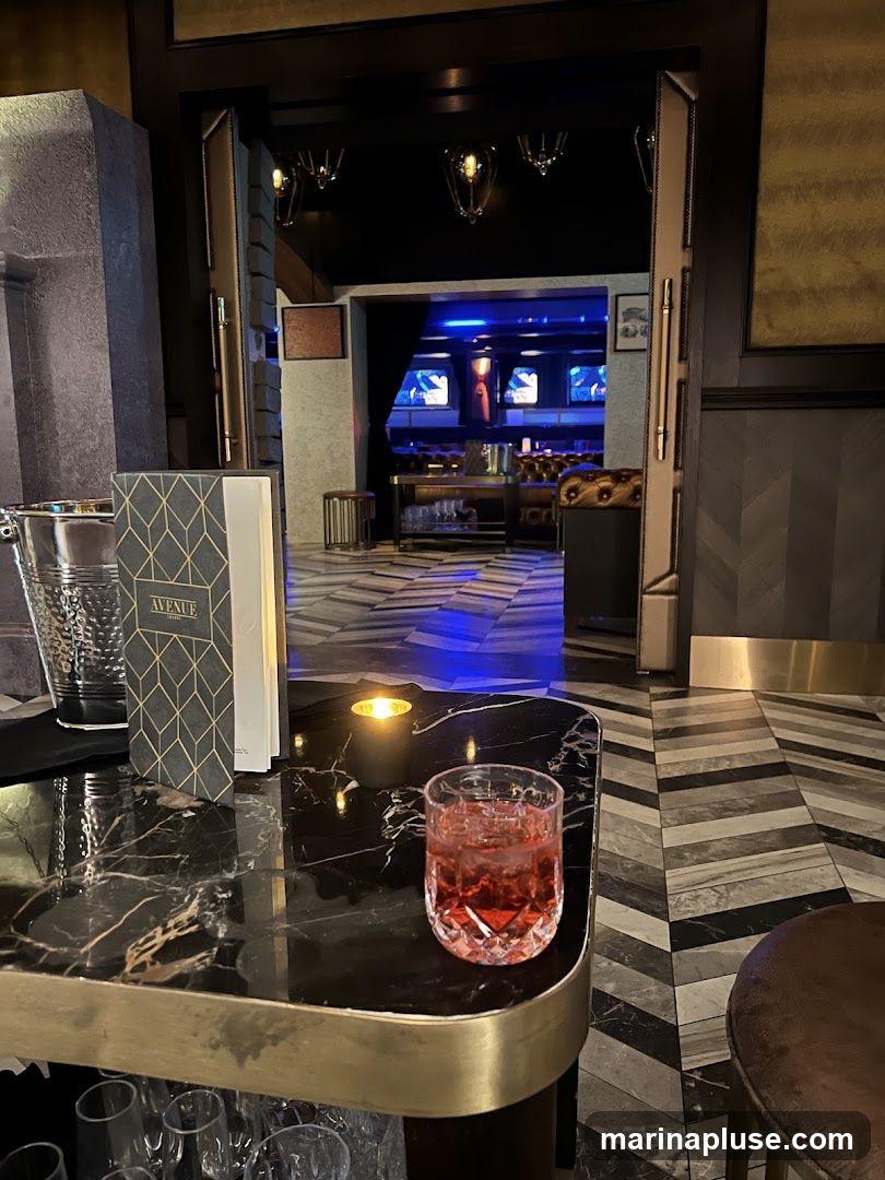 A dimly lit bar interior with a drink on a marble-topped table in the foreground. The drink is a reddish color in a faceted glass. A menu holder with the word "AVENUE" is also on the table. In the background, through an open doorway, a lounge area with plush seating and screens displaying content is visible, illuminated by blue lights.