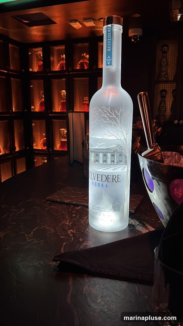 A bottle of Belvedere Vodka is illuminated on a dark, marble-patterned bar. Behind the bottle, shelves are stocked with various bottles, many of which appear to contain red liquid and are lit from within. To the right of the vodka bottle, an ice bucket with purple accents holds ice and tongs. A dark napkin and a menu are also visible on the bar.