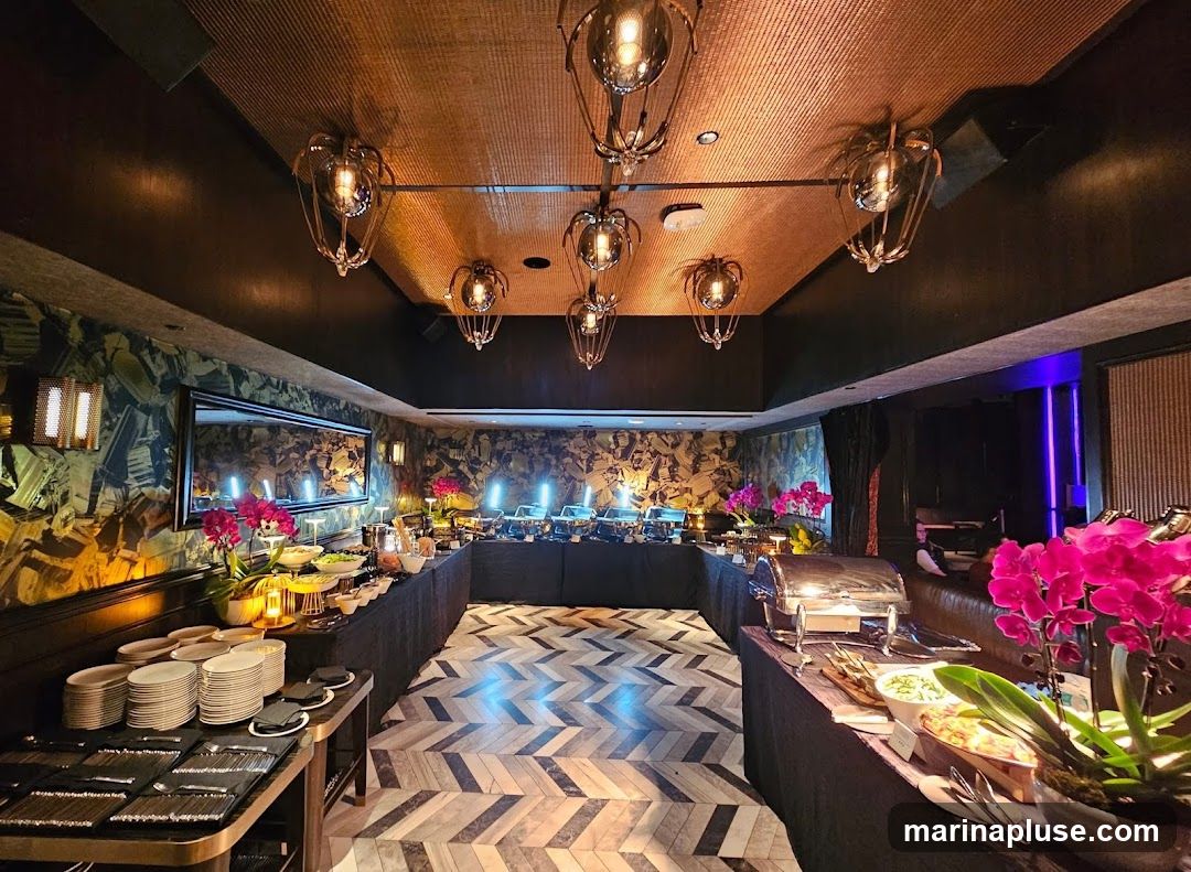 A long buffet table is set up in a room with ornate wallpaper and decorative lighting fixtures. The buffet features multiple chafing dishes, serving bowls, and stacks of plates and cutlery. Pink orchids are placed as floral arrangements. The floor has a distinctive herringbone pattern.