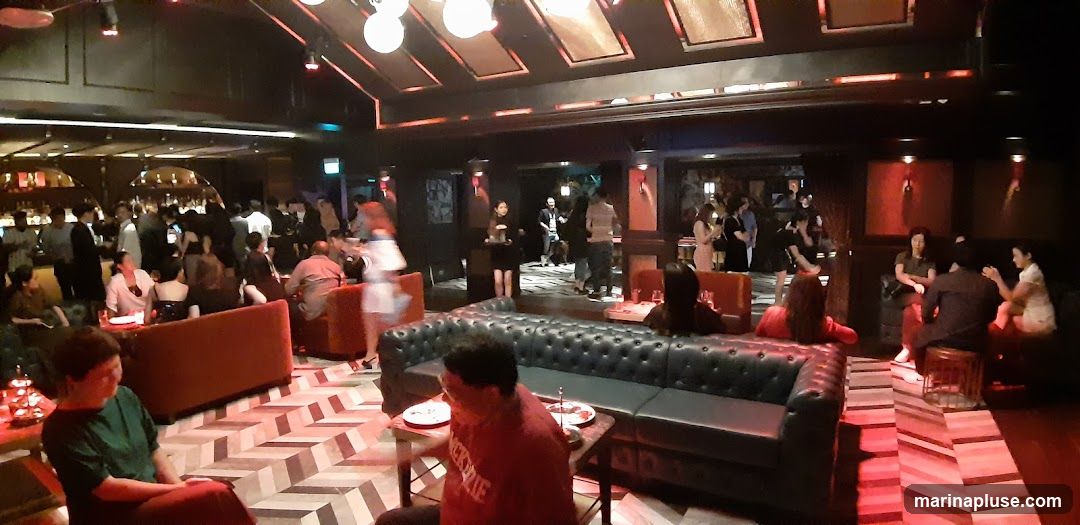 A dimly lit, upscale bar interior with numerous patrons socializing. The space features plush seating, including dark leather sofas and red upholstered chairs, arranged around tables. A long bar area is visible in the background, with bottles and glassware displayed. The ceiling has decorative lighting fixtures. The floor has a geometric pattern.