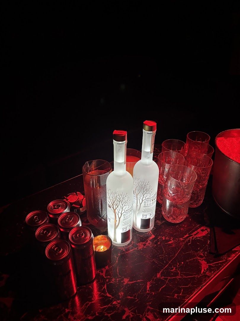 Two bottles of Grey Goose vodka are illuminated on a dark, marble-patterned bar. Several cans of a red beverage are arranged to the left, along with a small lit candle. Multiple glasses are stacked to the right, and a bucket filled with ice and red material is visible at the far right. The scene is lit with red ambient lighting, creating a moody atmosphere.
