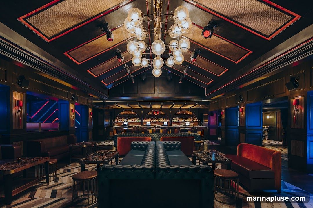 A dimly lit, opulent lounge area with plush seating, marble-topped tables, and a long bar in the background. The ceiling features a dramatic chandelier and spotlights, with red and blue accent lighting throughout the space. The decor suggests a high-end establishment, possibly a nightclub or upscale bar.