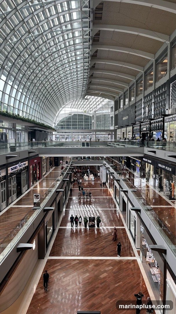 A wide-angle view of the interior of a large, multi-level shopping mall. The architecture features a distinctive curved glass roof and a spacious, polished wooden floor. Several shops with illuminated storefronts are visible on multiple levels, with shoppers walking along the corridors. The overall impression is one of a bright, contemporary, and upscale retail environment.