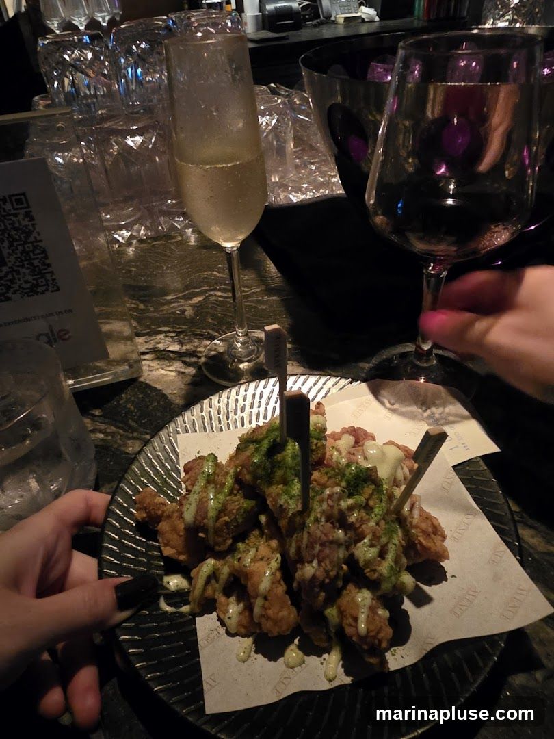 A plate of fried chicken pieces, drizzled with white sauce and sprinkled with green seasoning, is served on a textured plate with paper lining. Two glasses, one with champagne and another with red wine, are visible on the bar counter. Several empty glasses and an ice bucket are also present. A QR code is visible on a small sign.