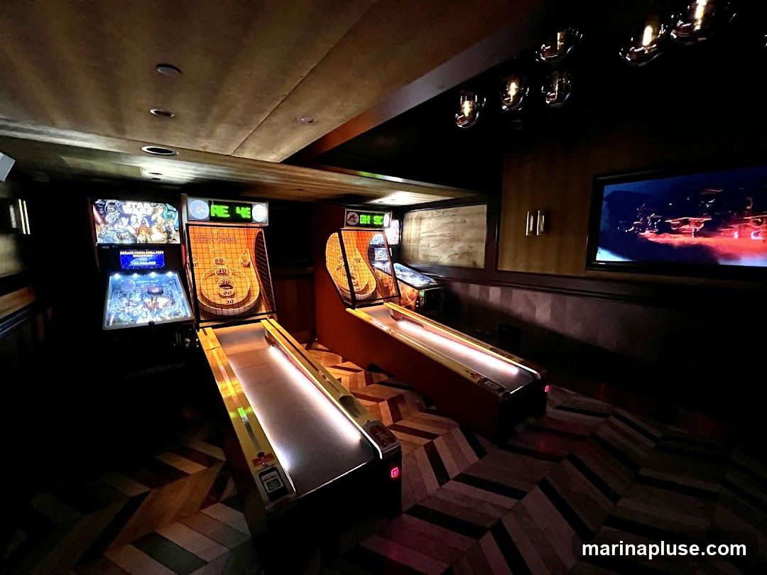 A dimly lit room with several arcade games, including a pinball machine and Skee-Ball machines. A large screen is mounted on the wall, displaying a scene with red and blue lights. The floor has a herringbone pattern.
