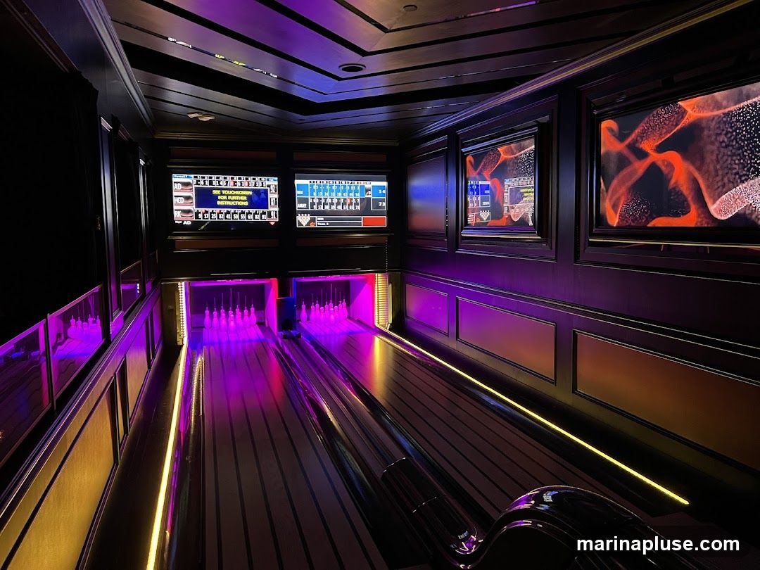 A dimly lit bowling alley with purple and yellow accent lighting. Two bowling lanes are visible, with pins set up at the end of each. Multiple screens display scores and graphics. Decorative framed artwork adorns the walls.