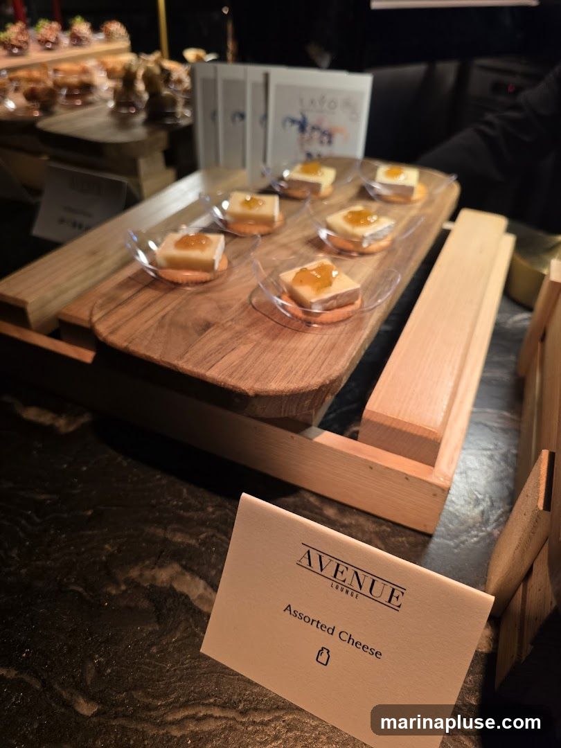 A close-up view of a cheese platter with small appetizers. The appetizers consist of a square of cheese topped with a dollop of orange jam, served on a cracker with a smear of orange sauce. They are arranged on a wooden board with a light-colored wooden stand. In the foreground, a small card reads "AVENUE LOUNGE" and "Assorted Cheese" with a small icon below. In the background, there are more appetizers on platters and some branded boxes.