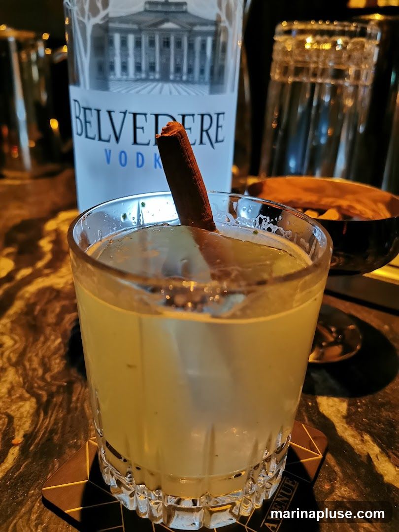 A close-up shot of a cocktail in a glass, garnished with a cinnamon stick. A bottle of Belvedere Vodka is visible in the background, along with stacked glasses and a bowl of snacks. The drink appears to be a light yellow color and is served on a patterned coaster.