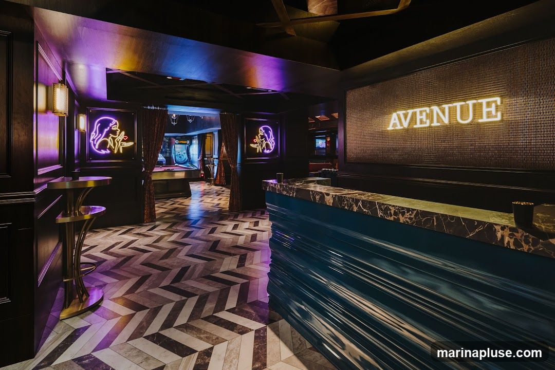 The interior of a dimly lit bar features a marble-topped counter with a teal, fluted base. Behind the counter, a large wall display has the word "AVENUE" illuminated in bright yellow neon. To the left, a dark wood paneled wall has a neon sign of a woman blowing a kiss in purple. Further into the bar, a pool table and arcade games are visible, with red velvet curtains framing the entrance to that area. The floor has a distinctive herringbone pattern of grey and beige tiles.