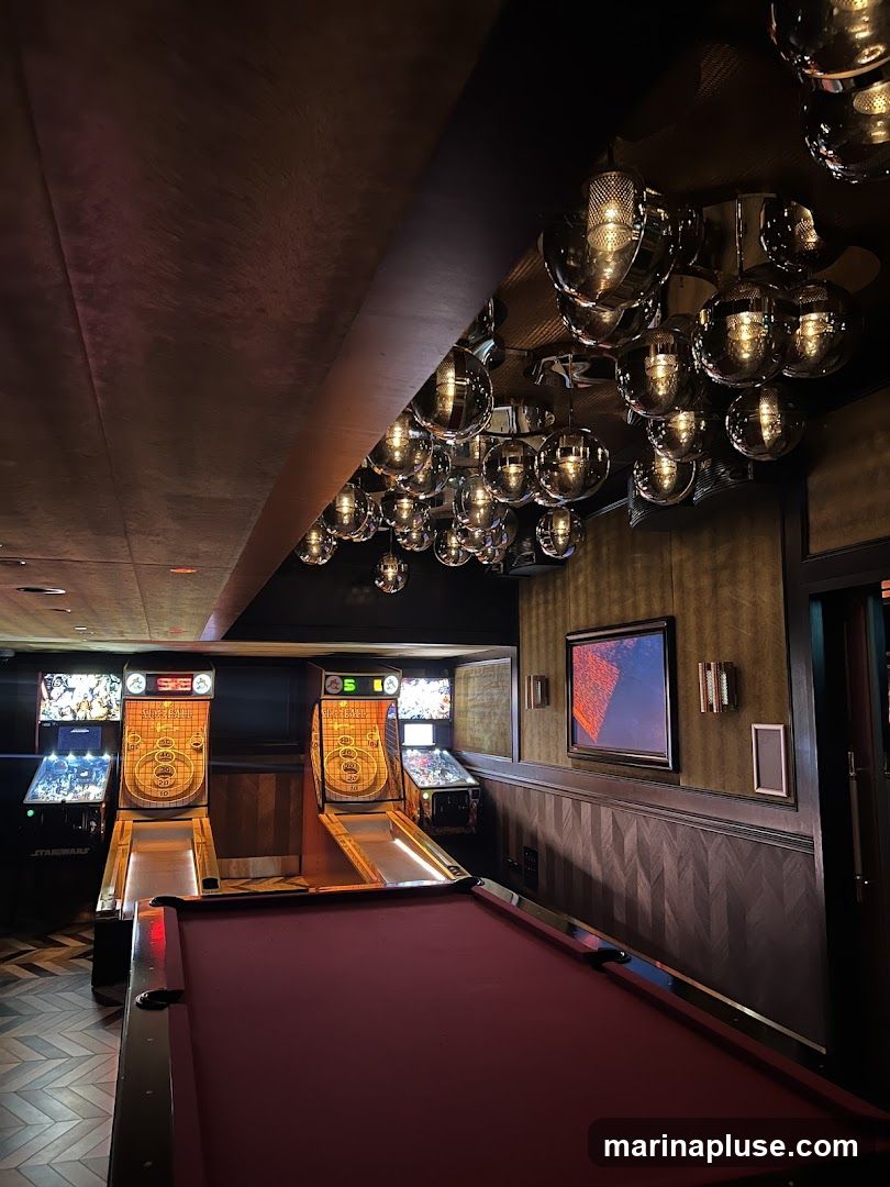 A dimly lit room features a pool table in the foreground. Behind it are several arcade games, including Skee-Ball machines and pinball machines. The ceiling is adorned with numerous reflective spherical light fixtures, creating a dynamic and moody ambiance. A framed artwork hangs on the wall.