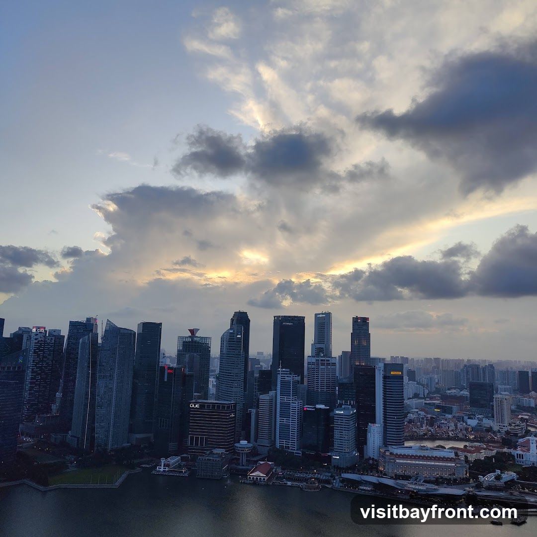 A panoramic view of a modern cityscape with numerous skyscrapers, seen from a high vantage point. The sky is filled with dramatic clouds, with rays of sunlight breaking through, suggesting either sunrise or sunset. A body of water, likely a bay or river, is visible in the foreground, reflecting the city lights. The architecture is diverse, with many tall, glass-fronted buildings characteristic of a major financial or business district.