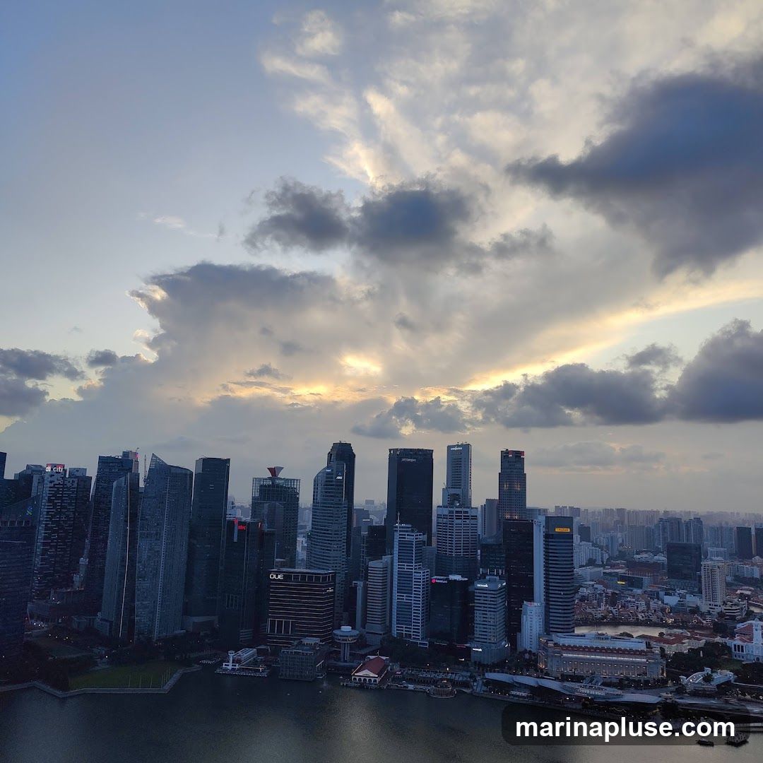 A panoramic view of a modern cityscape with numerous skyscrapers, seen from a high vantage point. The sky is filled with dramatic clouds, with rays of sunlight breaking through, suggesting either sunrise or sunset. A body of water, likely a bay or river, is visible in the foreground, reflecting the city lights. The architecture is diverse, with many tall, glass-fronted buildings characteristic of a major financial or business district.
