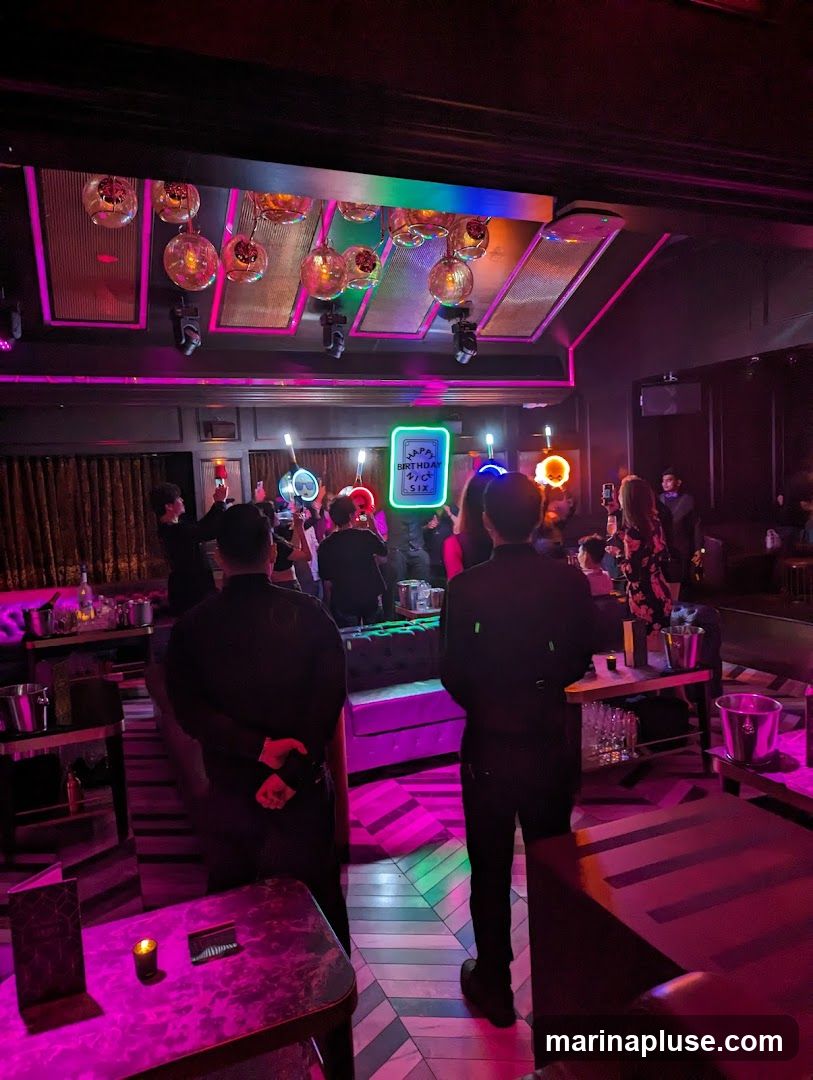 The image depicts the interior of a nightclub or lounge, illuminated by vibrant pink and purple lighting. Several patrons are visible, some holding up sparklers or glow sticks, suggesting a celebration. A neon sign in the center reads "HAPPY BIRTHDAY TO SIX". Staff members in black uniforms are also present, attending to guests. The decor includes ornate light fixtures hanging from the ceiling and plush seating areas.