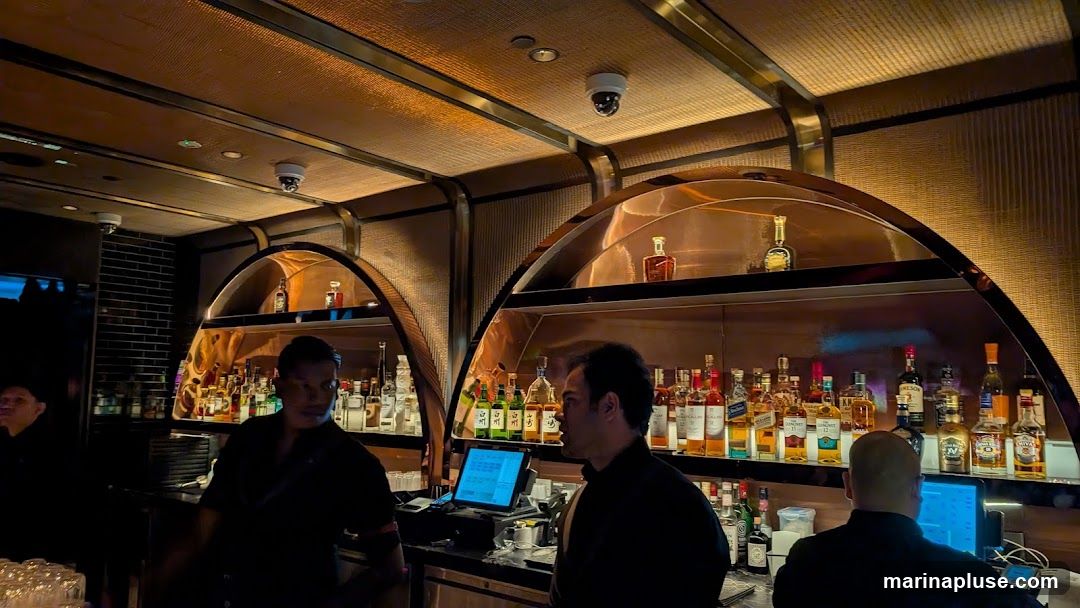 A dimly lit bar interior with a long counter stocked with numerous bottles of alcohol on shelves. Several bartenders are visible behind the counter, some interacting with customers or operating a computer system. The decor features warm lighting and textured walls.
