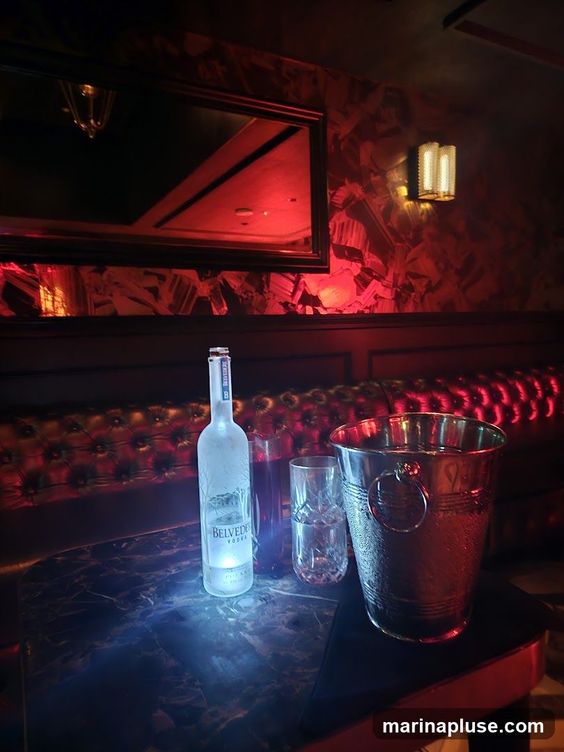 A bottle of Belvedere Vodka, two glasses, and a silver ice bucket are arranged on a marble-topped table. The scene is illuminated by red lighting, casting a dramatic glow on the surroundings. A tufted leather banquette is visible in the background, along with a large mirror and a wall sconce.