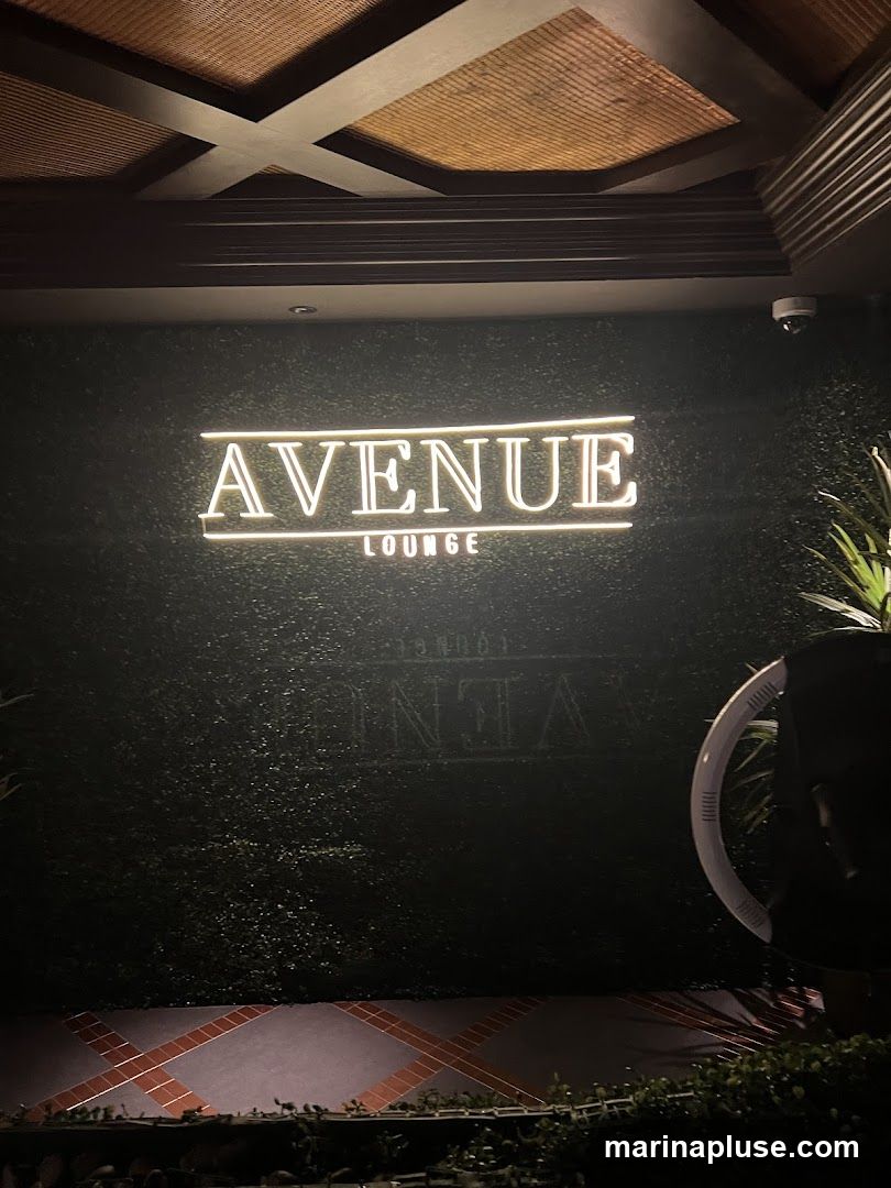 A neon sign that reads "AVENUE LOUNGE" is mounted on a dark green, textured wall. The sign is illuminated with white light. Below the sign, a faint, larger text is visible on the wall, but it is not legible. The ceiling above has a decorative pattern with wooden beams and woven panels. The lower part of the image shows a tiled floor with a geometric pattern and some greenery.