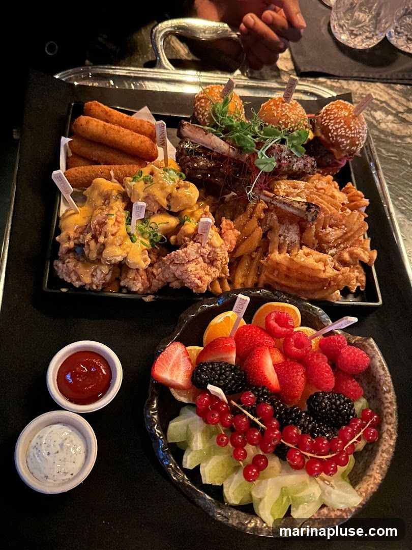 A large platter filled with various appetizers, including fried chicken, sliders, ribs, and fries. A side dish of fruit salad with berries and citrus fruits is also visible, along with two dipping sauces.