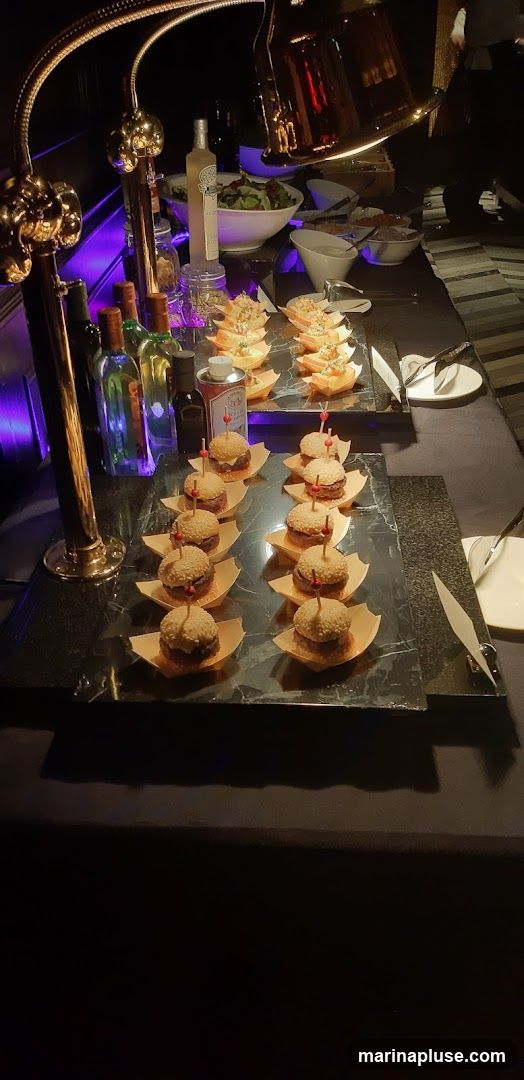 A buffet table is set with various food items, including mini sliders and appetizers. There are bottles of wine and other beverages on the table, as well as bowls of salad and dips. The lighting is dim, with spotlights illuminating the food.