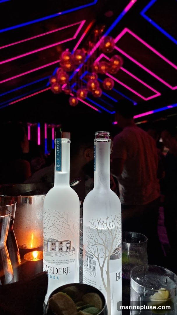 Two frosted bottles of Belvedere Vodka are displayed on a table in a dimly lit establishment. The bottles feature an etching of a manor house and a tree. In the background, neon lights in pink and blue create a vibrant atmosphere, and blurred figures of people can be seen. A lit candle and glasses with ice and lime wedges are also visible on the table.