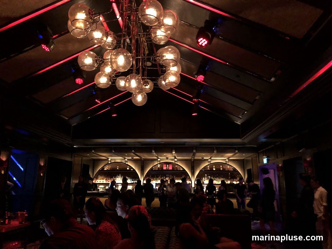 The image shows the interior of a dimly lit bar with a large, ornate chandelier hanging from the ceiling. Red and blue lights are visible, casting a moody ambiance. Several people are seated and standing, with a bar visible in the background.