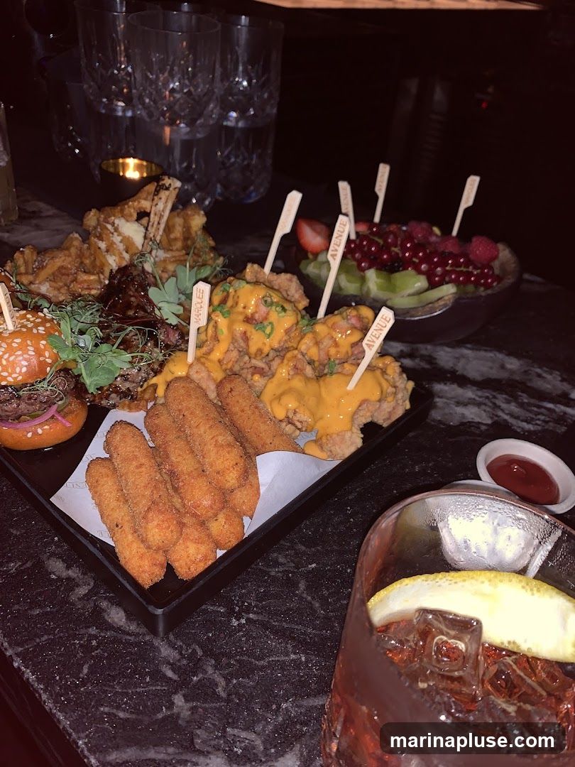 A platter of assorted appetizers including a mini burger, ribs, fried chicken, mozzarella sticks, and a fruit salad. A cocktail with a lemon slice and a small dish of dipping sauce are also visible. Wooden skewers with the word "AVENUE" are stuck into some of the food items.